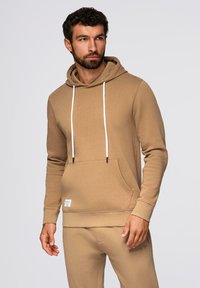 Homme aux cheveux foncés et à la barbe portant un sweat à capuche beige avec des cordons blancs et un pantalon beige assorti, regardant sur le côté.