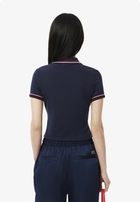 Navy polo shirt with red and white collar accents, fitted design, smooth texture, paired with high-waisted navy trousers and a red bag.