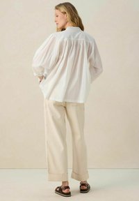 White, oversized shirt with a gathered back, paired with loose beige trousers and brown sandals, featuring a rolled cuff design.
