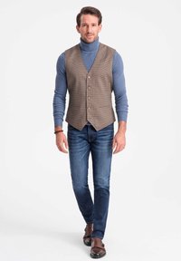 Braunes Hahnentrittmuster-Westen mit fünf Knöpfen über einem blauen Rollkragenpullover, kombiniert mit blauen Jeans und braunen Doppel-Monk-Schnürschuhen.