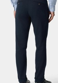 Granatowe chinosy o dopasowanym kroju cechują się gładką teksturą, tylkimi kieszeniami z guzikami oraz podwiniętymi mankietami. Materiał wydaje się lekki i elegancki.