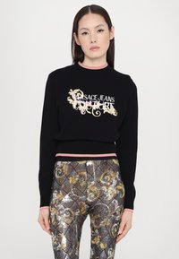 Svart strikket genser med brodert "Versace Jeans Couture"-logo; ribbestrikket halslinning og hem med rosa detaljer, kombinert med glanset mønstrede leggings.