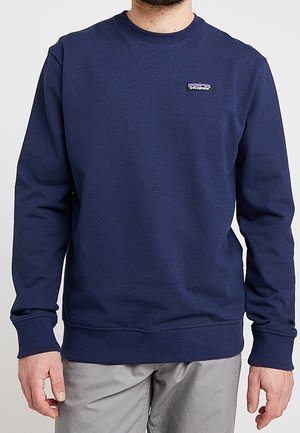 Mężczyzna noszący prostą granatową bluzę z okrągłym dekoltem z małym logo Patagonia po lewej stronie klatki piersiowej, w połączeniu z jasnoszarymi spodniami.