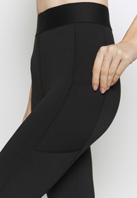 Leggings noirs avec une taille haute, fabriqués en matériau extensible. Comprend des coutures visibles et une poche latérale pour le rangement.