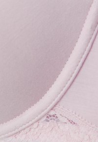 Vaaleanpunainen rintaliivi, jossa on sileä kangas, pitsireunus ja pyöristetty kaula-aukko. Ominaisuuksiin kuuluu pehmeä rakenne ja huomaamaton ommel reunoilla.