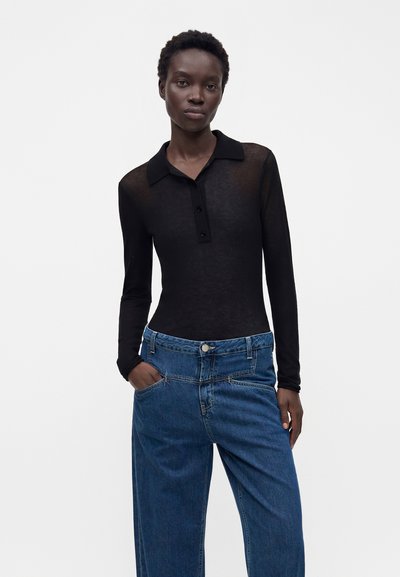 Femme portant un polo noir ajusté à manches longues et un jean bleu ample, avec une main dans la poche, debout devant un fond uni.