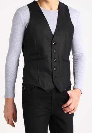 Mann trägt eine eng anliegende schwarze Weste mit Knöpfen über einem hellgrauen Langarmhemd und schwarzen Hosen, eine Hand ruht in der Nähe der Tasche.