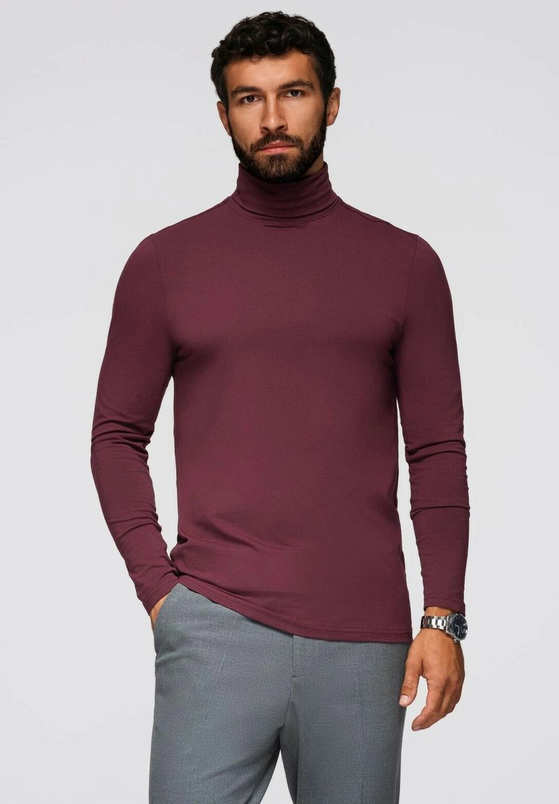 Vinröd turtleneck-tröja i mjukt material, långa ärmar och en figurnära design. I kombination med ljusgrå byxor, vilket ger en avslappnad look.