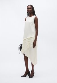 Rochie crem asimetrică, cu partea superioară netedă și detalii cu franjuri la tiv. Poartă pantofi negri ascuțiți și o geantă mică neagră.