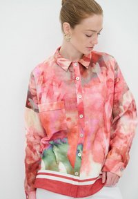 Camicia a pulsante trasparente e colorata con un motivo floreale astratto in rosa, rosso e verde, dotata di taschino sul petto e orlo a strisce.
