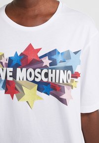 Bílý tričko s krátkým rukávem, které má velký grafický nápis "LOVE MOSCHINO" spolu s multibarevnými třpytivými hvězdami a výbuchy v různých barvách.