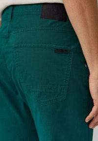 Teal katoenen broeken met een achterzak voorzien van een zwart label. De stof is soepel met een lichte glans en zichtbare stiksels.