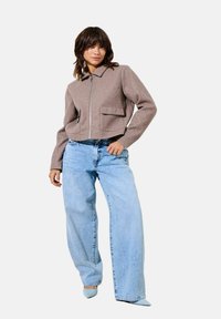 Korte bruine wollen jas met een rits, twee voorzakken en een kraag, gecombineerd met lichtblauwe wijde jeans en blauwe hakken.