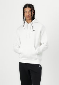 Felpa bianca Nike con tasca frontale, cappuccio regolabile e logo nero. Presenta maniche lunghe e una vestibilità comoda. Indossata con pantaloni neri.