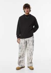 Zwarte lange mouwen shirt met een relaxte pasvorm, gecombineerd met gestreepte lichtgekleurde broek en tan schoenen. Accessoires zijn een zilveren kettinghalsband.