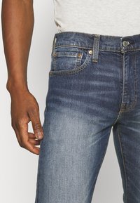 Tmavě modré džínové kalhoty se zúženým střihem, s pětiprvkovým designem kapes, žlutým stehem a mosazným kováním na pásku.