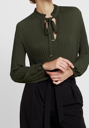 Frau trägt dunkelgrüne, plissierte Bluse mit Schleifenkragen und schwarze Hose mit hohem Bund, die Hände sanft auf der Taille verschränkt.