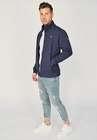 Granatowa bluza z zamkiem błyskawicznym z wysokim kołnierzem, o dopasowanym kroju, z logo na lewym rękawie. Noszona z jasnoniebieskimiciemnymi jeansami i białymi sneakersami.