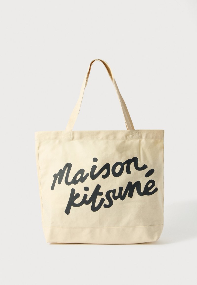 Plátěná taška v světle béžové barvě s černým ručně psaným textem "maison kitsuné." Má dva horní popruhy a obdélníkový tvar.