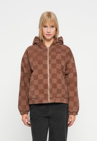 Donna bionda che indossa una giacca a zip con cappuccio a quadri marroni e pantaloni neri, in piedi su uno sfondo bianco.