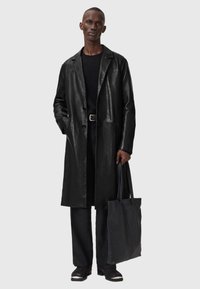 Cappotto lungo in pelle nera con scollo a colletto, due bottoni e tasche laterali. Abbinato a un maglione nero, pantaloni e una borsa tote nera con texture.