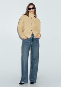 Beige ribbad cardigan med hög krage och svarta knappar, bärs över en svart-vit randig topp och ljusblå vida jeans.