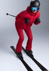 Piros, párnázott sídzseki, piros szűkített sí nadrág, fekete síbakancs és síléc, fekete kesztyű, valamint fekete sisak sötétített plexivel.