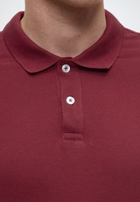 Burgundy polo shirt with a textured fabric, featuring a classic collar and three white buttons at the neck for fastening.