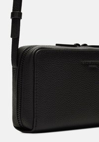 Schwarze Leder-Crossbody-Tasche mit strukturierter Oberfläche, rechteckiger Form, Reißverschluss und schlankem, verstellbarem Trageriemen. Markenlogo sichtbar.