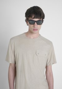 T-shirt beige a maniche corte con finitura testurizzata e tasca sul petto con due bottoni. Occhiali da sole rettangolari scuri con lenti a specchio.