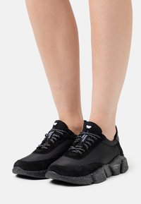 Pieds portant des baskets noires avec des semelles épaisses mouchetées et des lacets à motifs, présentés sur un fond blanc uni.