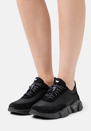 Pies con zapatillas negras de suela gruesa y moteada y cordones con patrón, mostrados sobre un fondo blanco liso.