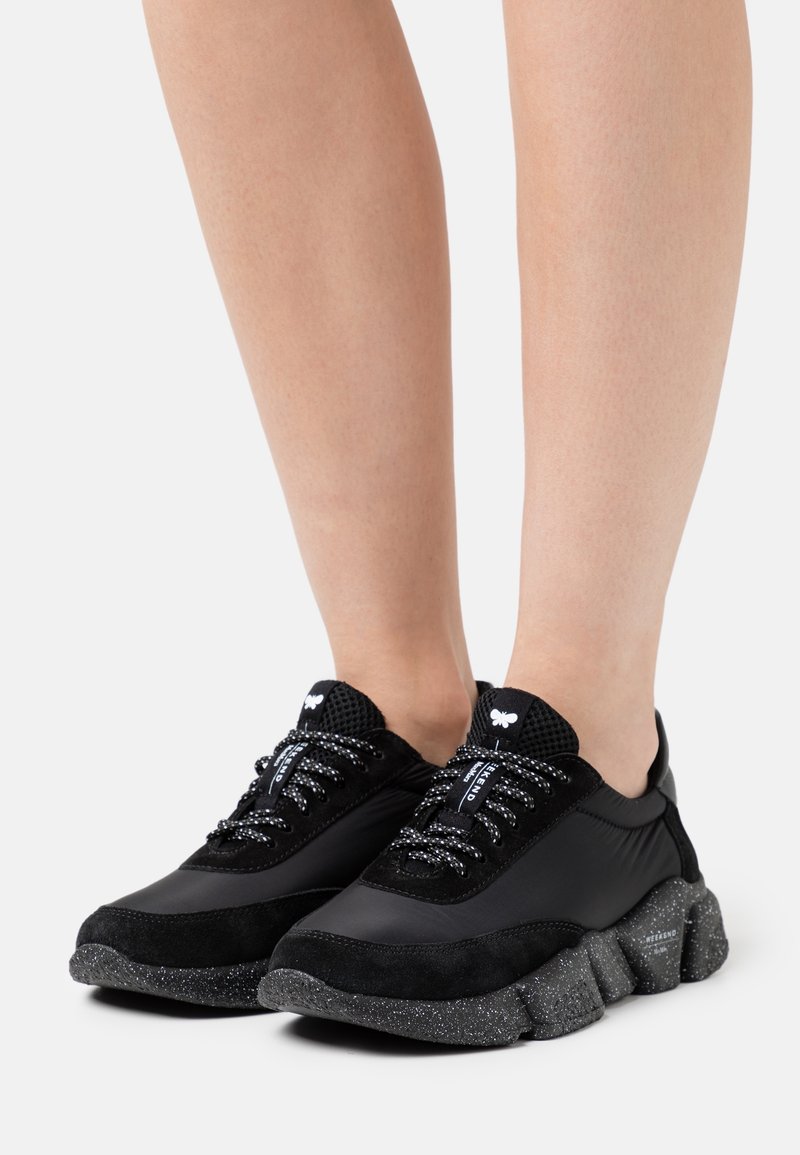 Pieds portant des baskets noires avec des semelles épaisses mouchetées et des lacets à motifs, présentés sur un fond blanc uni.