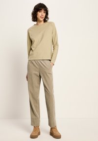 Frau steht in einem beigen Langarm-Shirt, hellbraunen Cordhosen mit Taschen und tanfarbenen Wildleder-Plattform-Ankle-Boots vor einem schlichten Hintergrund.