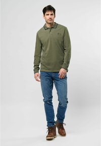 Žalia olive spalvos polo maikutė ilgomis rankovėmis su apykakle, su mažais logotipais. Suderinta su mėlynais džinsais ir rudais kuo užrišamais batais.