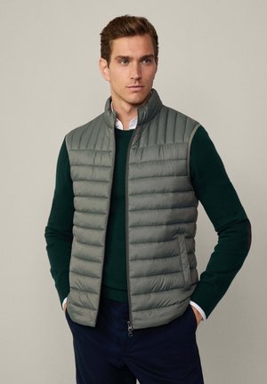 Graue Steppweste mit vertikaler Steppung, hohem Kragen und Reißverschluss. Über einem dunkelgrünen Pullover getragen, was den Lagenlook betont.