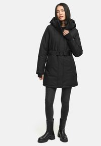Parka nera imbottita con vita con cintura, chiusura con zip e cappuccio regolabile. Presenta maniche lunghe e tasche laterali. Indossata con jeans neri e stivali.