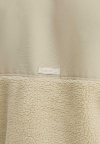 Pullover in pile color crema con una parte superiore liscia e una parte inferiore testurizzata. Presenta un piccolo etichetta bianca con la scritta "BLEND" sul lato.