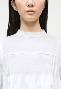 Γυναίκα με μακριά σκουρόχρωμα μαλλιά που φοράει λευκό μπλουζάκι με ψηλό γιακά και κάθετες πιέτες στο στήθος.