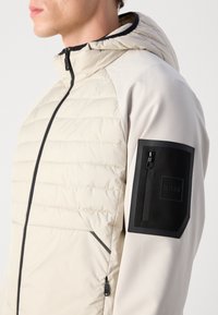 Beige puffer jacket with a hood, featuring a black zippered pocket on the sleeve and black zipper detailing. Smooth texture and modern design.