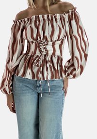 Bluzka odsłaniająca ramiona w kremowym kolorze z brązowymi falistymi wzorami, z talią ściągniętą sznurowaniem i bufiastymi rękawami. W połączeniu z jeansami.