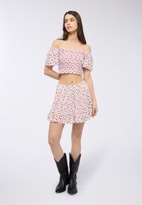 Top corto off-the-shoulder e gonna svasata di colore bianco con motivo floreale rosso. Stivali cowboy neri completano il look.