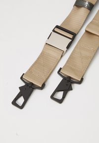 Sangles en nylon beige avec des clips en plastique noirs. Comprend une boucle en métal et des coutures horizontales pour une durabilité accrue.
