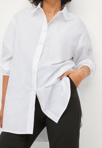 White button-up shirt made of lightweight fabric, featuring a classic collar, long sleeves, and a relaxed fit with side slits.