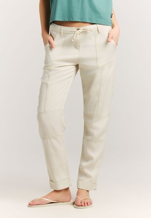 Kvinde iført beige snørebuks med op-rullede manchetter og beige klip-klappere, stående med hænderne i lommerne mod en ensfarvet baggrund.