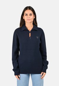 Pull bleu marine avec col haut, fermeture moitié éclair et un onglet en cuir. Présente un petit logo brodé sur la poitrine. Texture tricotée douce.