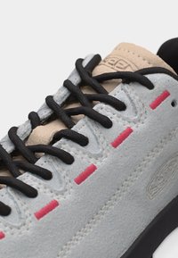 Κοντινό πλάνο γκρι σουέντ αθλητικού παπουτσιού με μαύρα κορδόνια, μπεζ γλώσσα και κόκκινα ραμμένα διακοσμητικά κατά μήκος των οπών για τα κορδόνια και της πλευρικής επιφάνειας.