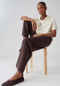 Vrouw zittend op een houten kruk met één been omhoog, gekleed in een crèmekleurige korte mouw trui, donkerbruine broek en bijpassende platte schoenen.