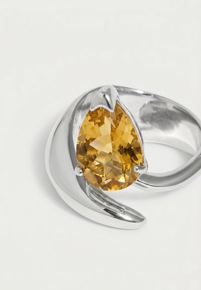 Anillo de plata con una piedra preciosa facetada dorado-amarilla en forma de lágrima, montada en un diseño metálico moderno y curvado sobre un fondo blanco.