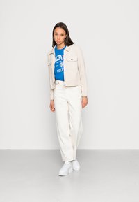 Chaqueta de pana beige con dos bolsillos frontales, camiseta gráfica azul, jeans de pierna ancha blancos y zapatillas de plataforma blancas. Look limpio y casual.